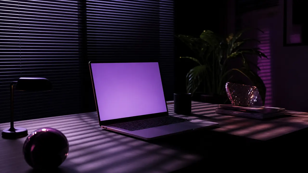 A laptop glowing violet in a dark room with blind shadows